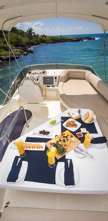 Luxury yacht breakfast table with orange juice, fruit platter and pastries, overlooking turquoise sea and tropical rocky coastline under a blue sky