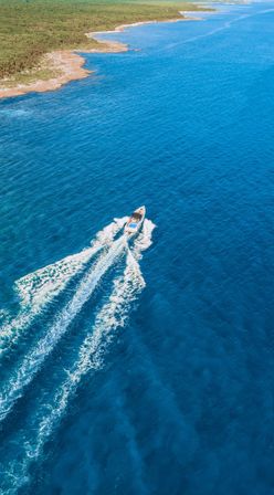 Aerial view of a motorboat zipping across clear turquoise ocean, leaving twin white wakes near a rocky tropical coastline
