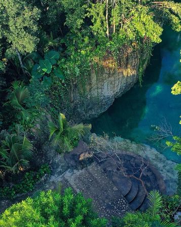 Aerial view of a tropical cliffside and turquoise lagoon framed by dense jungle and palm trees, with stone steps descending to emerald-blue water.