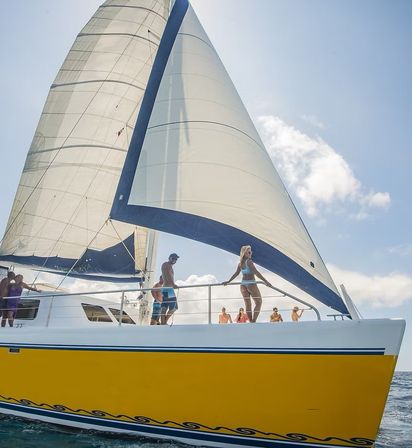 Yellow-hulled catamaran with large white sails and people in swimwear on deck, cruising on blue ocean under a sunny sky