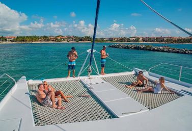 Couple and friends relaxing on a white catamaran's front trampoline nets and deck in clear turquoise tropical water near a palm‑lined resort shoreline under a sunny blue sky.