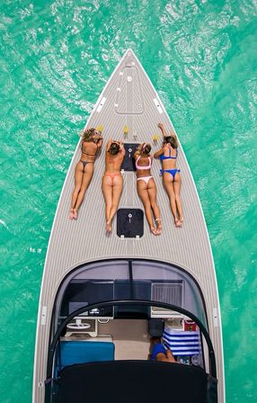 Aerial view of four friends in colorful bikinis sunbathing on the bow of a yacht over crystal turquoise tropical water.