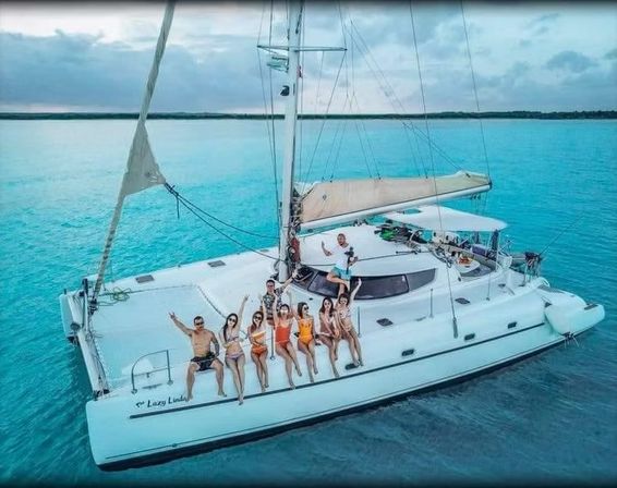Group of friends in swimsuits lounging on the bow of a white catamaran anchored in clear turquoise tropical waters under a partly cloudy sky