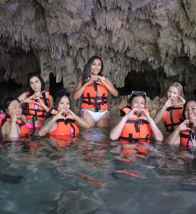 Group of people in orange life jackets waist-deep in a limestone cave pool, smiling and forming heart shapes with their hands beneath dramatic stalactites
