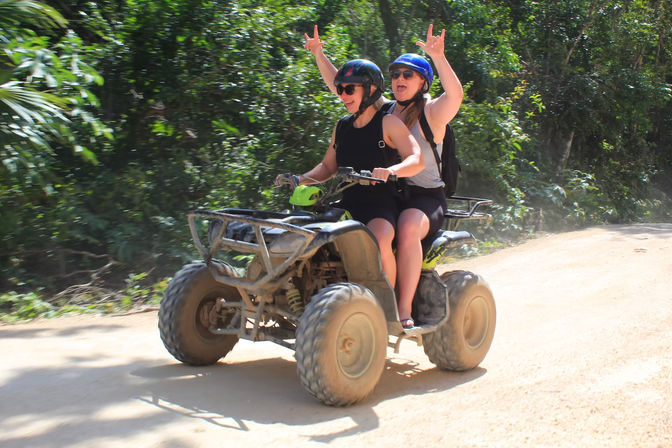 Two riders on an ATV on a dusty dirt trail through lush tropical forest, passenger cheering with hands raised for an off‑road adventure