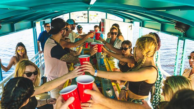 Group of young adults toasting with red plastic cups inside a covered party boat on a sunny summer day, sunglasses and casual clothes with water visible through the windows.