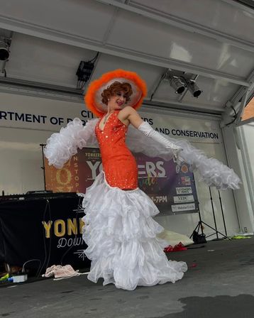Drag performer in an orange sequined gown and matching wide-brim hat with long white ruffled skirt and gloves, striking a pose on an outdoor city festival stage