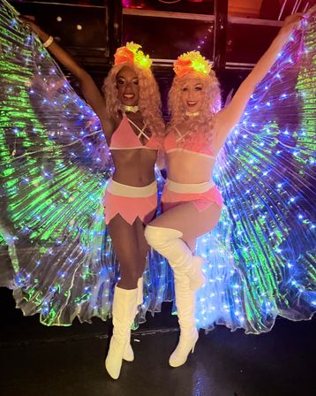 Two performers in matching pink fairy costumes with glowing LED wings, flower headpieces and white thigh-high boots posing with arms raised on a colorful nightclub dance floor — vibrant nightlife party vibe.