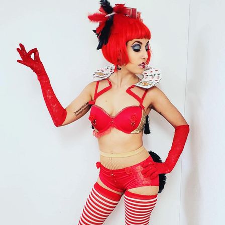 Cabaret/cosplay performer in a playing-card inspired red costume — bright red bob wig with feathered headpiece, red bra and high-waist shorts, striped red-and-white thigh-highs, long red gloves and a collar made of playing cards, posing against a white background.