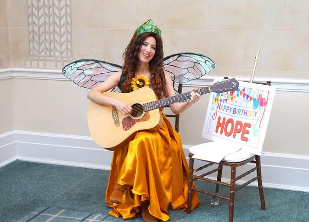 Performer in a fairy costume with iridescent wings, green cap and sunflower brooch, wearing a golden satin dress and playing an acoustic guitar while seated in an indoor event room beside a colorful 'Happy Birthday' sign.