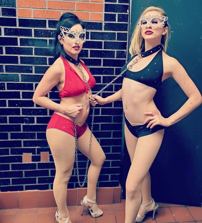 Two masked performers in masquerade-style masks and matching collar chokers, dressed in red and black bikini costumes with fishnet tights and high heels, linked by a silver chain and posing against a dark brick wall.