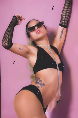 Glam fashion model in a black bikini and cat-eye sunglasses striking a dramatic pose against a pink studio backdrop, wearing fishnet arm sleeves, a long rhinestone necklace and visible tattoos — edgy retro studio look.