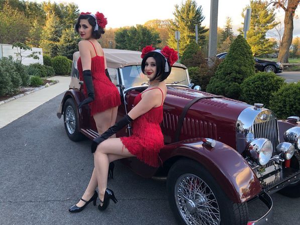 Two performers in red 1920s-style flapper dresses and black gloves posing on a maroon vintage convertible parked on a suburban street at golden hour, surrounded by trees and shrubs.