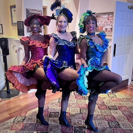 Three can-can cabaret dancers in red, blue, and green ruffled costumes with feathered hats and fishnet stockings, kicking together at an indoor event venue