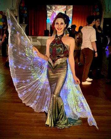 Smiling performer in a sequined mermaid gown and glowing LED wings posing on a wooden dance floor at an indoor party, with colorful balloons and a stage with red curtains in the background.
