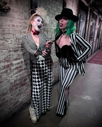 Two costumed performers in black-and-white striped outfits pose against a textured brick hallway: one in white face paint, feathered headpiece and harlequin skirt, the other with green hair, black hat and striped pants playfully pulling a tie.