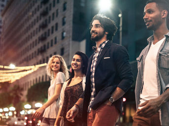 Four friends smiling as they walk down a lively downtown street at night, urban nightlife with glowing string lights and building facades in the background.