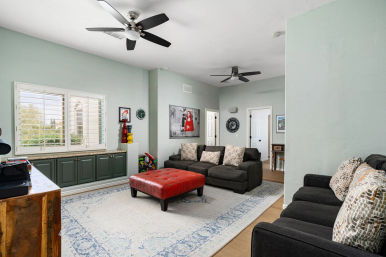 Bright family living room with mint-green walls, shuttered window over green built-in cabinets, dark gray sofas with patterned pillows, red tufted leather ottoman on a blue-accent area rug, two ceiling fans and wall art.