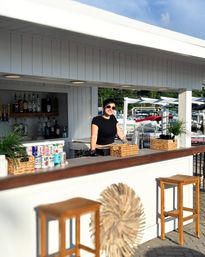 Smiling bartender in sunglasses behind a white dockside bar at a sunny marina, with bottles and canned beers on the counter, wicker baskets, wooden stools and boats in the background.