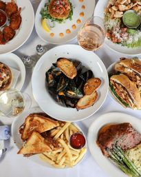 Top-down restaurant spread: steamed mussels with toasted bread, club sandwich and fries with ketchup, steak with asparagus and mash, grilled chicken salad, crab cake, fried shrimp and glasses of wine.