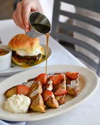 Hand drizzling maple syrup from a metal pitcher over powdered French toast slices topped with fresh strawberries and a dollop of whipped cream on a brunch plate.
