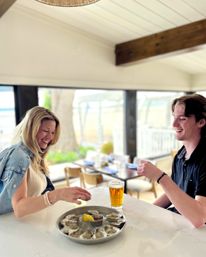 Two people laughing at a bright waterfront seafood bar sharing an oyster platter on ice with lemon and cocktail sauce and a pint of beer, coastal restaurant dining