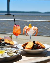 Seaside seafood spread on a wooden table: chilled shrimp cocktail with lemon, fresh oysters on the half shell, mussels with garlic toast, and a bright orange summer cocktail, with a sunny waterfront and railing in the background.