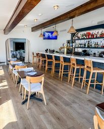 Bright coastal-style restaurant interior with a long white bar and brass taps, row of wooden barstools, wicker pendant lights, exposed wooden beams, and sunlit dining tables set with napkins