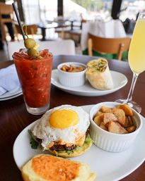 Brunch at a bright cafe — egg-topped burger with runny yolk, toasted bun with spicy spread, roasted breakfast potatoes, Bloody Mary with olives and a mimosa.