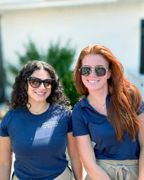 Two smiling restaurant servers in sunglasses and navy tops with beige aprons posing on a sunny outdoor dining patio, one with curly dark hair and one with long red hair.
