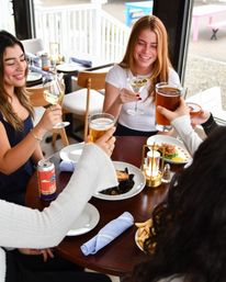 Friends toasting on a sunlit restaurant patio with wine, a martini with olives, pints of beer and shared plates of mussels and fries