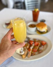 Hand holding a chilled mimosa flute over a white-tablecloth brunch spread — French toast topped with strawberries and whipped cream, a breakfast sandwich and a Bloody Mary blurred in the background
