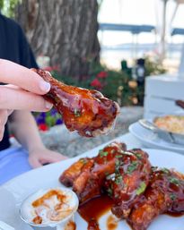 Hand holding a sticky BBQ-glazed chicken drumette above a plate of saucy wings with ranch dip, garnished with herbs on a waterfront patio with blurred trees and dock in the background.