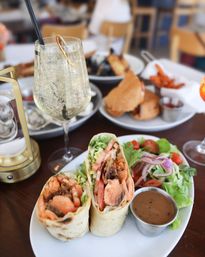 Two salmon wraps with lettuce, tomato and crispy bacon on a plate with mixed greens and dipping sauce, bubbly white wine cocktail on a restaurant table, casual brunch scene
