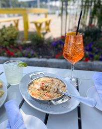 Cheesy baked lobster mac and cheese in a small skillet on a white outdoor patio table, paired with a sparkling orange spritz and a lime cocktail, yellow picnic benches and summer flowers blurred in the background.