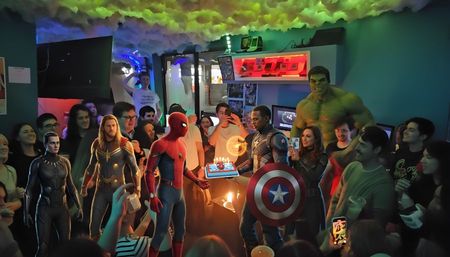 Neon-lit indoor gaming lounge birthday party with a crowd gathered around a superhero-themed cake held by Spider-Man and Captain America figures, flanked by Thor, Hulk and Black Panther cutouts under colorful ceiling lights.