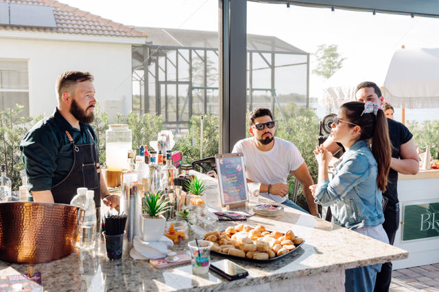 Sunny outdoor patio bar scene: bearded bartender in an apron chatting with casually dressed guests over a granite counter filled with bottles, a drink menu sign, colorful mixers, and a tray of bite-sized pastries — relaxed lakeside/backyard vibe.