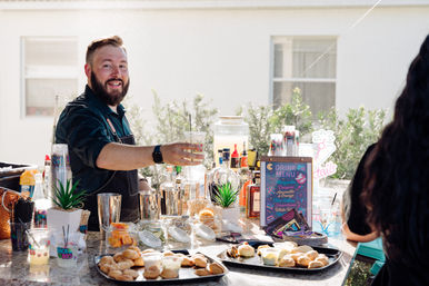 Smiling bartender offers a drink at a sunlit backyard patio bar setup with bottles, mixers, a colorful drink-menu sign, and trays of pastries and garnishes on the counter.