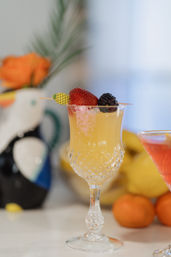 Cut-crystal goblet holding a pale orange citrus cocktail, topped with a strawberry and blackberry on a playful pineapple pick, on a bright tropical-style brunch table with blurred fruit in the background.