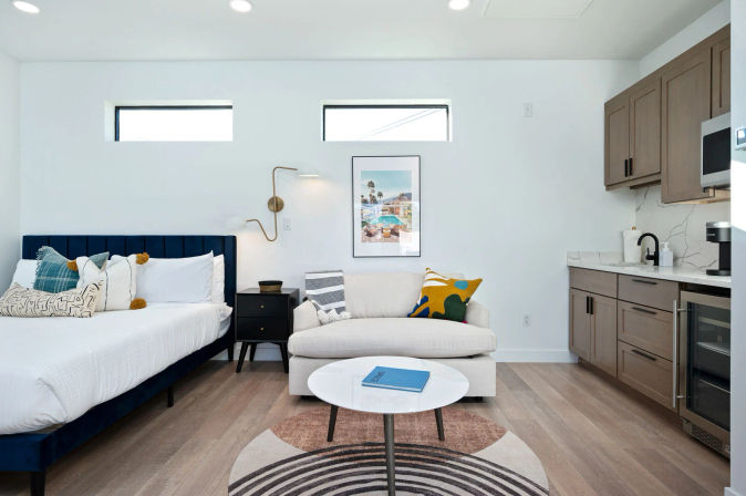 Bright modern studio apartment with navy-upholstered bed, white loveseat with colorful pillows, round marble-top coffee table on a geometric rug, and compact wood-tone kitchenette beneath two horizontal clerestory windows.
