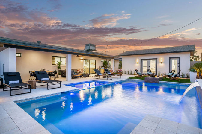 Inviting modern backyard with an illuminated blue swimming pool and waterfall feature, tiled patio lined with lounge chairs, outdoor seating and dining areas beside a single-story home under a colorful sunset sky