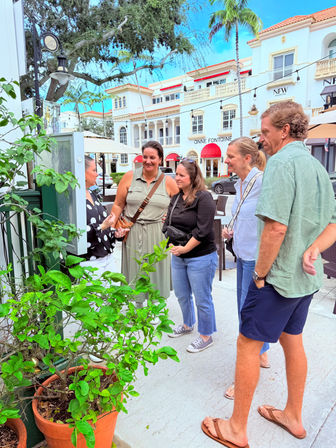 Five casually dressed adults smiling and chatting on a palm-lined coastal shopping street with white Mediterranean-style storefronts, red awnings, outdoor café seating and potted plants.