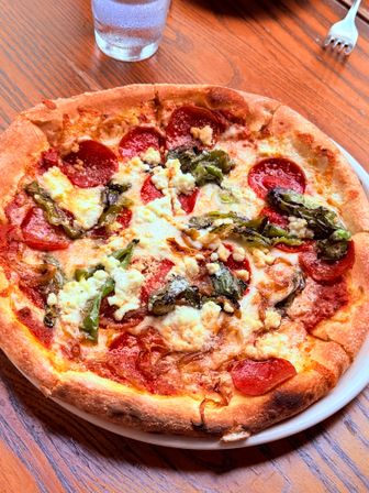 Mouthwatering thin‑crust pepperoni pizza with melted mozzarella, ricotta dollops, roasted green peppers and caramelized onions on a white plate on a wooden table with a chilled glass of water