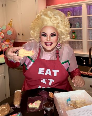 Drag performer with voluminous blonde wig and bold makeup wearing a red "EAT IT" apron in a bright home kitchen, smiling and holding a decorated sugar cookie over a counter with baking and cookie-decorating supplies.
