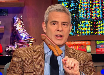 Gray-haired TV talk show host in a plaid jacket on a colorful studio set holding a novelty corn dog on a stick
