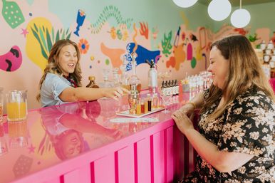 Two women at a bright pink bar sampling a cocktail tasting flight of small bottles and mixers with glassware and a colorful mural wall in the background.