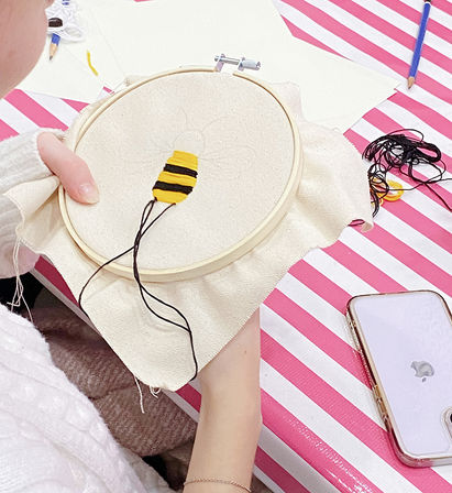 Close-up of a hand embroidery hoop with a yellow-and-black bee body being stitched, black floss trailing across cream fabric on a pink-and-white striped table, with a smartphone and threads nearby.