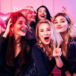 Five women laughing and posing together at a bachelorette-style party, wearing small veils and making a peace sign in front of a pink backdrop with balloons.