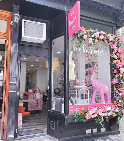 City fashion boutique storefront with black trim and floral garland, pink elephant window display, mannequin in chevron dress, sewing machine and open door to a cozy sewing studio.