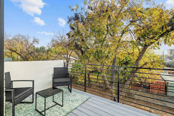 Rooftop balcony deck with two black woven chairs and a small metal table on a green patterned rug, modern metal railing overlooking trees with yellow autumn leaves and nearby residential rooftops.